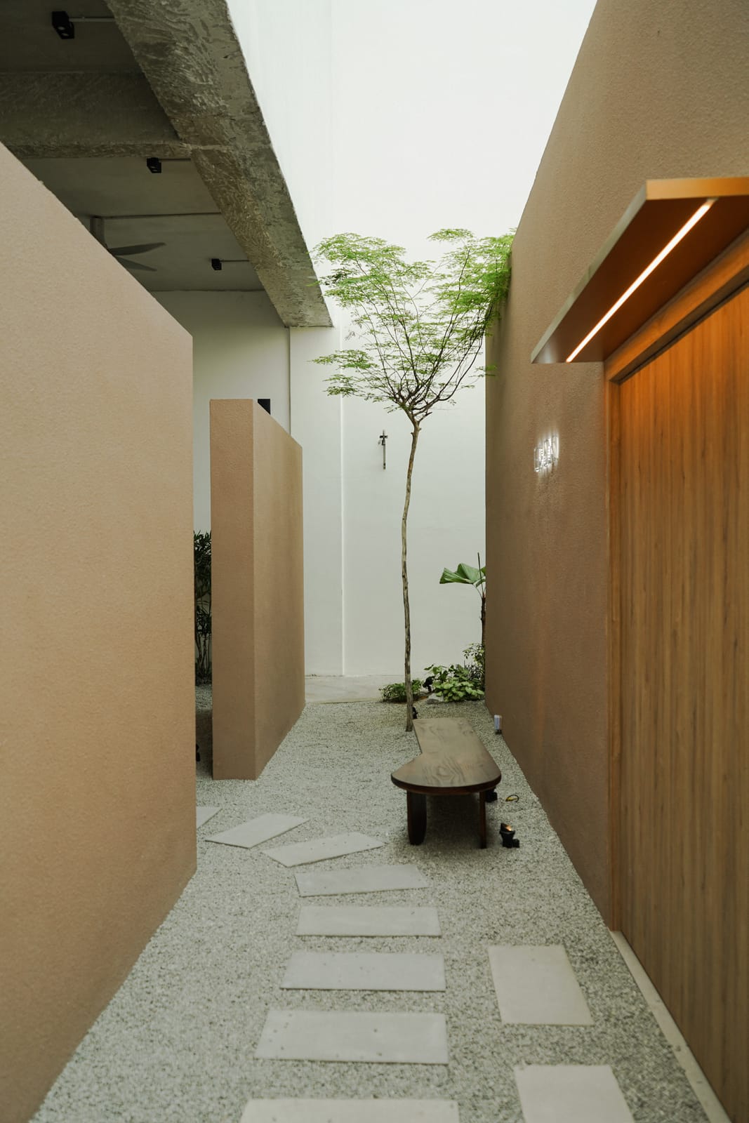 Eden's entrance path: stepping stones across white gravel between clay-coloured walls, a wooden bench and a young tree under an open skylight